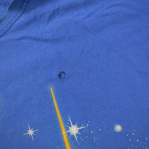 Pu&ograve; includere: Una maglietta blu con un piccolo buco nel tessuto. La maglietta ha una grafica sbiadita di una stella gialla e stelle bianche su uno sfondo blu.