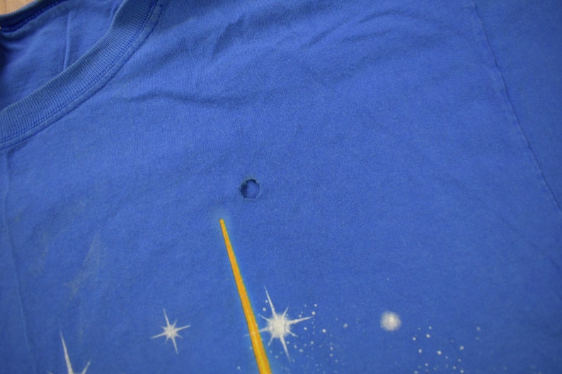 Pu&ograve; includere: Una maglietta blu con un piccolo buco nel tessuto. La maglietta ha una grafica sbiadita di una stella gialla e stelle bianche su uno sfondo blu.
