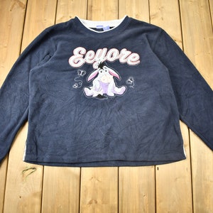 May include: A dark blue long-sleeved sweatshirt with a white inner collar. The front of the sweatshirt features a cartoon image of Eeyore from Winnie the Pooh, with the word "Eeyore" in pink and white letters above the image.