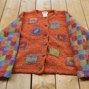 May include: A textured orange button-down cardigan featuring long sleeves with a multi-coloured checkered pattern in shades of blue, green, purple, and red. The front of the garment displays several small square patches with embroidered nature motifs.