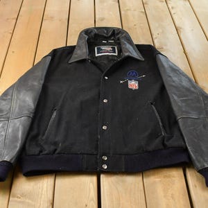 以下が含まれることがあります： ウール製のボディとレザー製の袖が特徴の黒いバーシティジャケット。レザーの襟、金属製のスナップボタン、リブ編みの袖口が付いています。胸元にはNFLのロゴと文字Mが入った円形のグラフィックが刺繍されています。