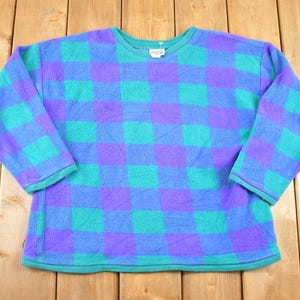 May include: A vintage fleece pullover in a vibrant checkerboard pattern of blue, teal, and purple. The long-sleeved top has a crew neck and teal trim. A tag is visible on the neckline. This retro style is perfect for casual wear.