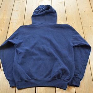 K&ouml;nnte beinhalten: Ein marineblauer Kapuzenpullover, von hinten gesehen, mit Kordelzugkapuze und gerippten B&uuml;ndchen und Saum. Der Pullover ist aus einem weichen, leicht abgenutzten Stoff gefertigt und liegt auf einer hellen Holzoberfl&auml;che.