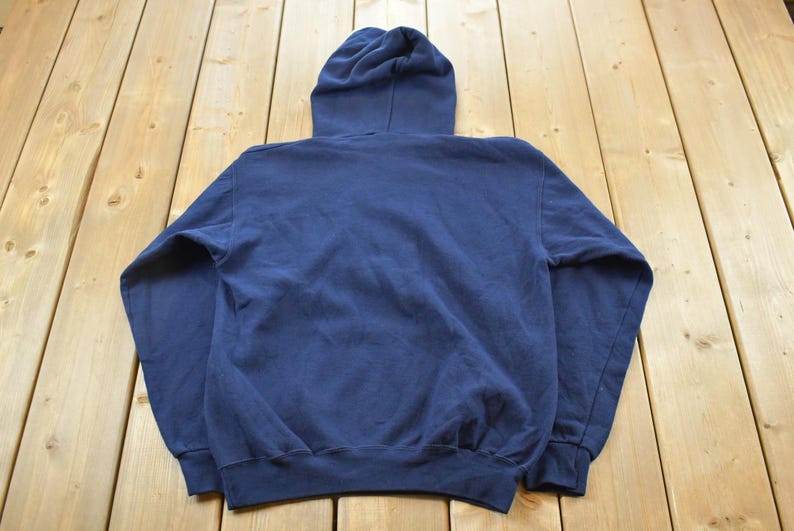 K&ouml;nnte beinhalten: Ein marineblauer Kapuzenpullover, von hinten gesehen, mit Kordelzugkapuze und gerippten B&uuml;ndchen und Saum. Der Pullover ist aus einem weichen, leicht abgenutzten Stoff gefertigt und liegt auf einer hellen Holzoberfl&auml;che.