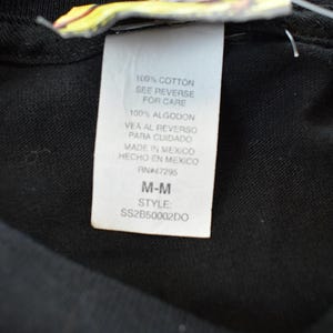 May include: A close-up of a black garment with a white label. The label states the item is 100% cotton, with care instructions. It also indicates the item was made in Mexico, size M-M, and style code SS2B50002DO.