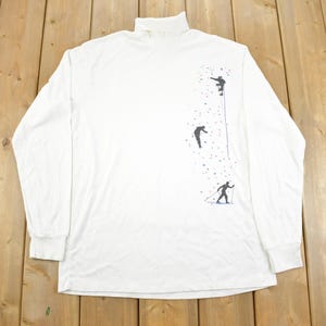 May include: White long-sleeve shirt with a turtleneck collar and a graphic print of three figures climbing a snowy mountain.
