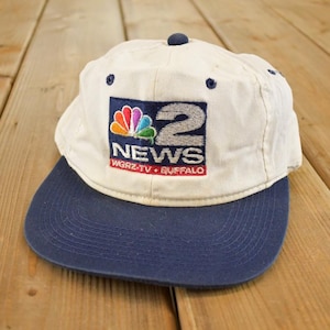 Op de afbeelding: Een vintage honkbalpet in wit en marineblauw. De pet heeft het "2 NEWS" logo met een kleurrijk ontwerp en de tekst "WGRZ-TV BUFFALO" in het rood. De pet is gemaakt van een zacht materiaal en heeft een gebogen klep.