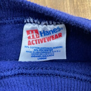 Op de afbeelding: Een blauw Hanes Activewear-label met witte tekst. Het label zegt "50% Katoen (Coton/Algodon) 50% Polyester Gemaakt in de VS Fabriqu&eacute; aux &Eacute;-U Hecho en los E.U.A. Grand/Grande Large".