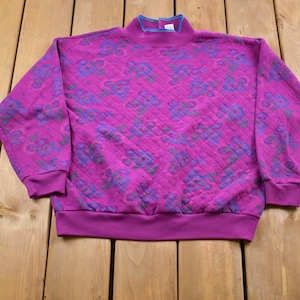May include: A pink sweatshirt with a quilted texture and a floral pattern in shades of purple, blue, and green. The sweatshirt has a mock neck and long sleeves.