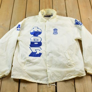 May include: White nylon jacket with blue lettering that reads "Alpha Beta Zeta" and a blue logo on the left chest.
