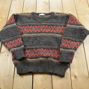 Peut inclure: Un pull en tricot gris anthracite avec un col rond et des manches longues. Le vêtement présente un motif horizontal répétitif de losanges rouges et de bandes géométriques complexes sur la poitrine, le dos et les manches, avec des bords-côtes.