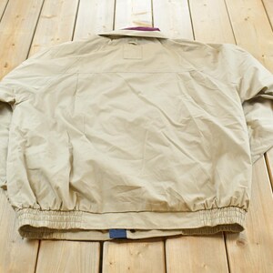 May include: Tan jacket with a purple collar and elastic cuffs. The jacket has a button closure and a small pocket on the back. The jacket is made of a lightweight material.