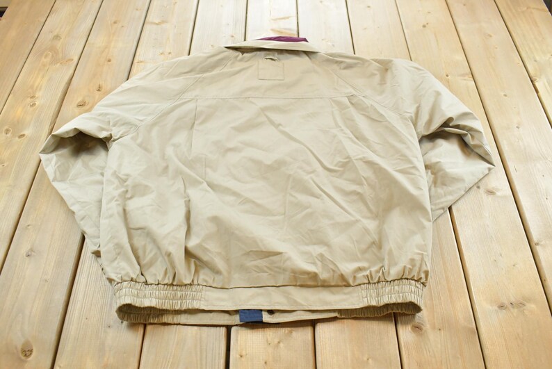 May include: Tan jacket with a purple collar and elastic cuffs. The jacket has a button closure and a small pocket on the back. The jacket is made of a lightweight material.