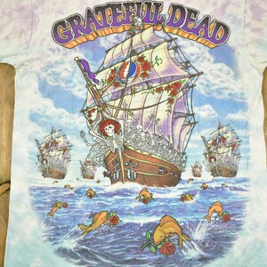 May include: A tie-dye t-shirt featuring a colourful image of a ship with skeletons on board sailing on a blue sea with fish and roses. The text "GRATEFUL DEAD" is printed in purple and yellow lettering at the top of the shirt.