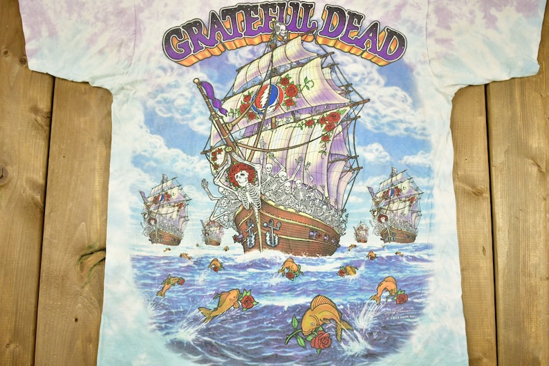 May include: A tie-dye t-shirt featuring a colourful image of a ship with skeletons on board sailing on a blue sea with fish and roses. The text "GRATEFUL DEAD" is printed in purple and yellow lettering at the top of the shirt.
