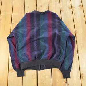 Puede incluir: Chaqueta bomber vintage con un estampado de rayas en tonos azules, verdes y burdeos. La chaqueta tiene cuello, pu&ntilde;os y dobladillo de canal&eacute; negro. La chaqueta est&aacute; sobre una superficie de madera.