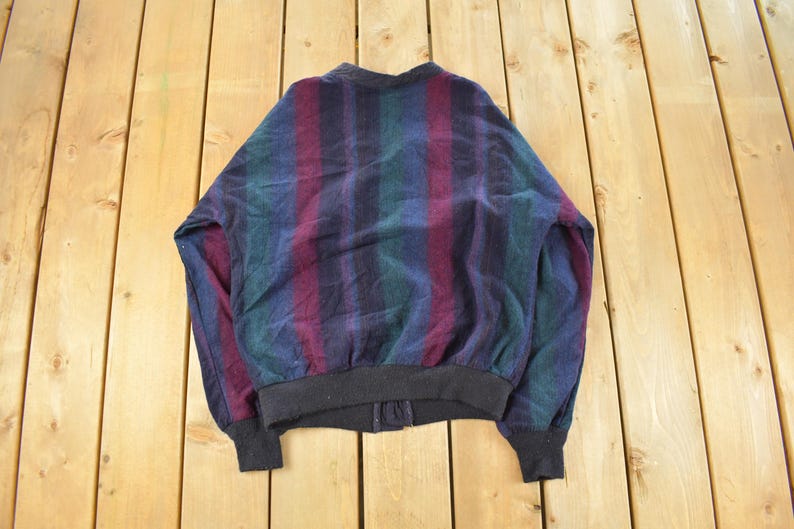 Puede incluir: Chaqueta bomber vintage con un estampado de rayas en tonos azules, verdes y burdeos. La chaqueta tiene cuello, pu&ntilde;os y dobladillo de canal&eacute; negro. La chaqueta est&aacute; sobre una superficie de madera.