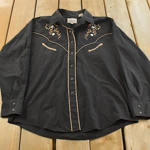 May include: Black long-sleeve button-down shirt with tan and white embroidered floral accents on the shoulders and chest. The shirt has tan piping along the front and a buttoned collar and cuffs. A vintage western-style shirt.
