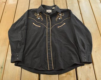 May include: Black long-sleeve button-down shirt with tan and white embroidered floral accents on the shoulders and chest. The shirt has tan piping along the front and a buttoned collar and cuffs. A vintage western-style shirt.