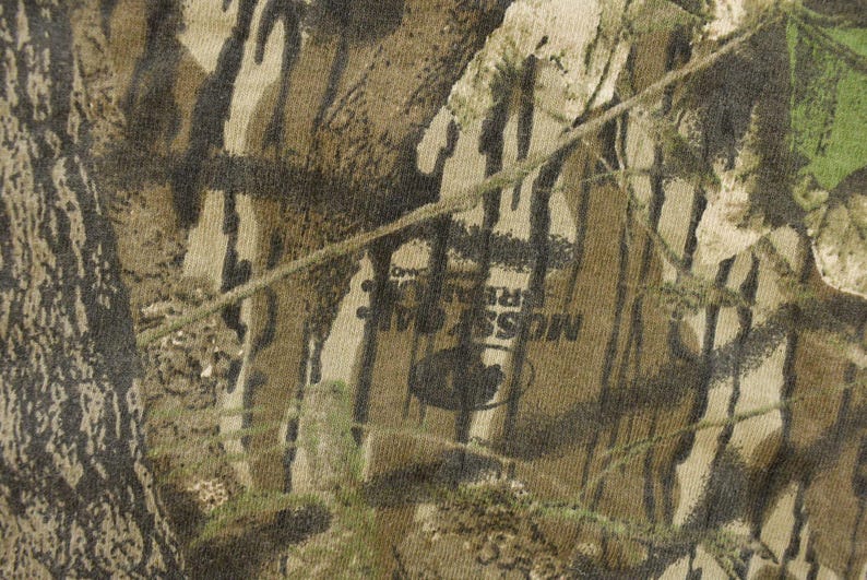 May include: Close-up of a camouflage pattern in shades of brown, green, and tan. The fabric has a textured appearance with the words "MOSS REAL TREE" visible. The design mimics a forest environment.