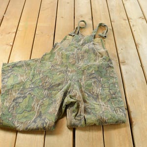 May include: Camouflage-patterned overalls with adjustable straps and multiple pockets. The green, brown, and tan colors create a woodland camouflage design. The overalls are made of a durable fabric, suitable for outdoor activities.