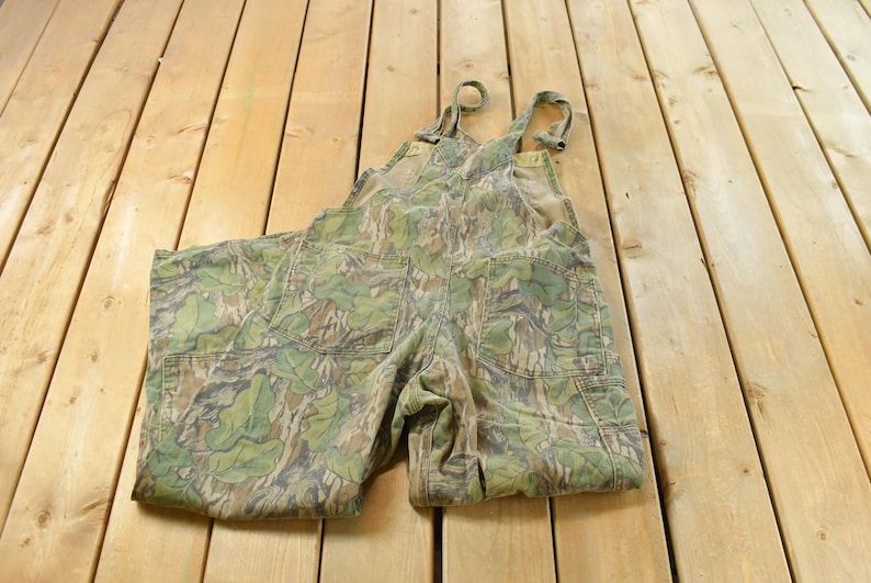 May include: Camouflage-patterned overalls with adjustable straps and multiple pockets. The green, brown, and tan colors create a woodland camouflage design. The overalls are made of a durable fabric, suitable for outdoor activities.