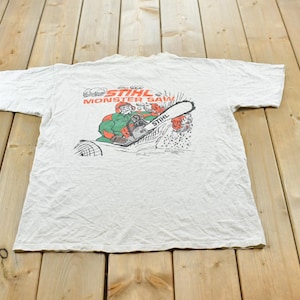 Op de afbeelding: Een lichtgrijs t-shirt met korte mouwen en een grafische print op de achterkant. Het ontwerp toont een stripfiguur die een kettingzaag bedient met de tekst STIHL MONSTER SAW in rood en zwart boven de illustratie.