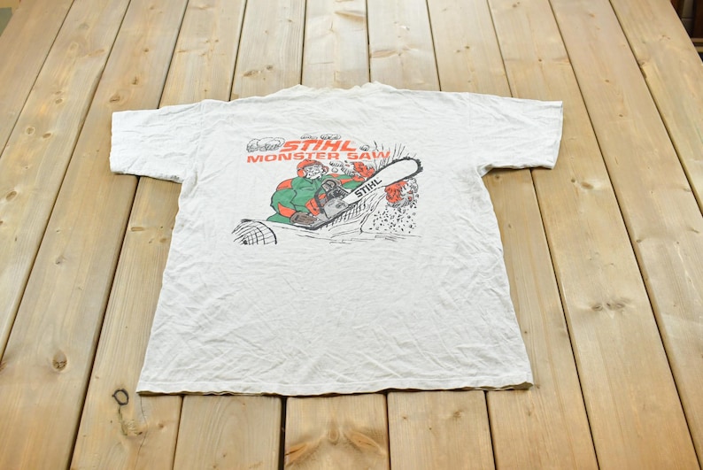Op de afbeelding: Een lichtgrijs t-shirt met korte mouwen en een grafische print op de achterkant. Het ontwerp toont een stripfiguur die een kettingzaag bedient met de tekst STIHL MONSTER SAW in rood en zwart boven de illustratie.