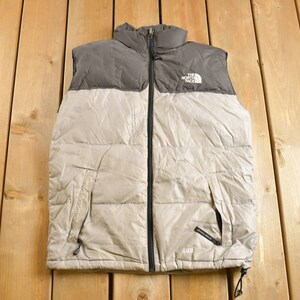 May include: A gray and light gray puffer vest with a black zipper and The North Face logo on the front. The vest has two zippered pockets on the sides and a drawstring at the bottom.