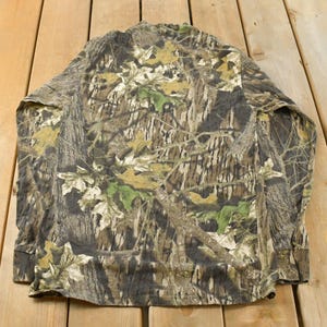 May include: A long-sleeved camouflage shirt in shades of brown, green, and beige. The shirt is displayed flat, revealing the back and sleeves. The fabric has a lightweight appearance, suitable for outdoor activities.