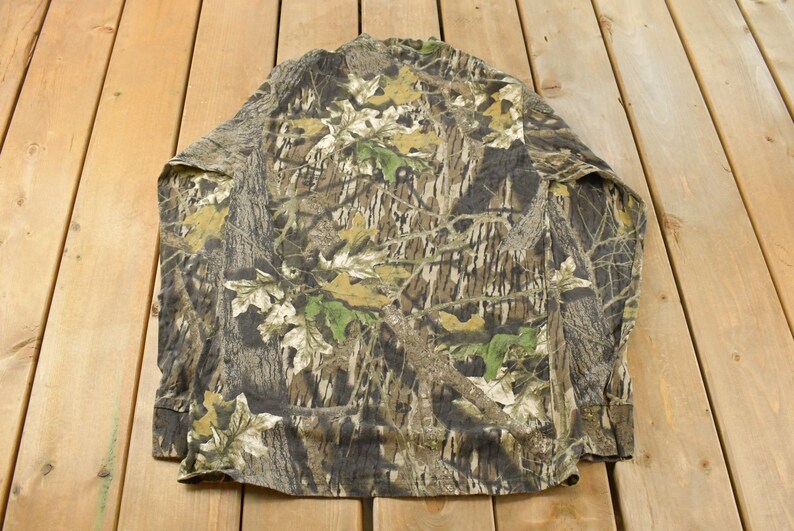 May include: A long-sleeved camouflage shirt in shades of brown, green, and beige. The shirt is displayed flat, revealing the back and sleeves. The fabric has a lightweight appearance, suitable for outdoor activities.