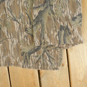 Op de afbeelding: Close-up van een shirt met camouflagepatroon. De stof heeft een mix van bruine, bruine en groene tinten, waardoor een natuurlijk, bosachtig ontwerp ontstaat. Het shirt heeft een zak en een ritssluiting.