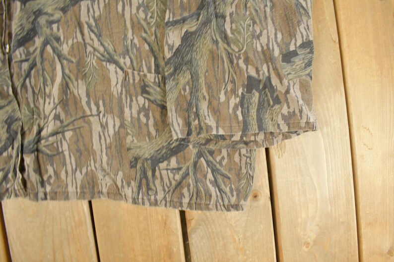 Op de afbeelding: Close-up van een shirt met camouflagepatroon. De stof heeft een mix van bruine, bruine en groene tinten, waardoor een natuurlijk, bosachtig ontwerp ontstaat. Het shirt heeft een zak en een ritssluiting.
