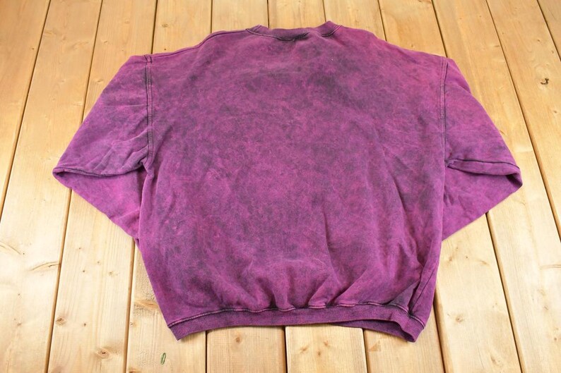 May include: A long-sleeve crewneck sweatshirt featuring a deep magenta and black acid-wash pattern. The garment has a ribbed collar, ribbed cuffs, and a ribbed hemline, showcasing a mottled, uneven color distribution across the entire fabric surface.