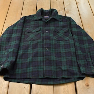 May include: A long-sleeve button-up shirt featuring a dark green and navy blue plaid pattern. The shirt includes a pointed collar, two chest pockets with buttoned flaps, and a full-length front button closure with matching dark buttons.