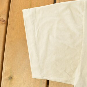 K&ouml;nnte beinhalten: Nahaufnahme eines cremefarbenen T-Shirt-&Auml;rmels. Der Stoff ist leicht zerknittert und der &Auml;rmel ist vor einem Holzgrund platziert. Das Shirt scheint aus einem weichen, leichten Material zu sein.