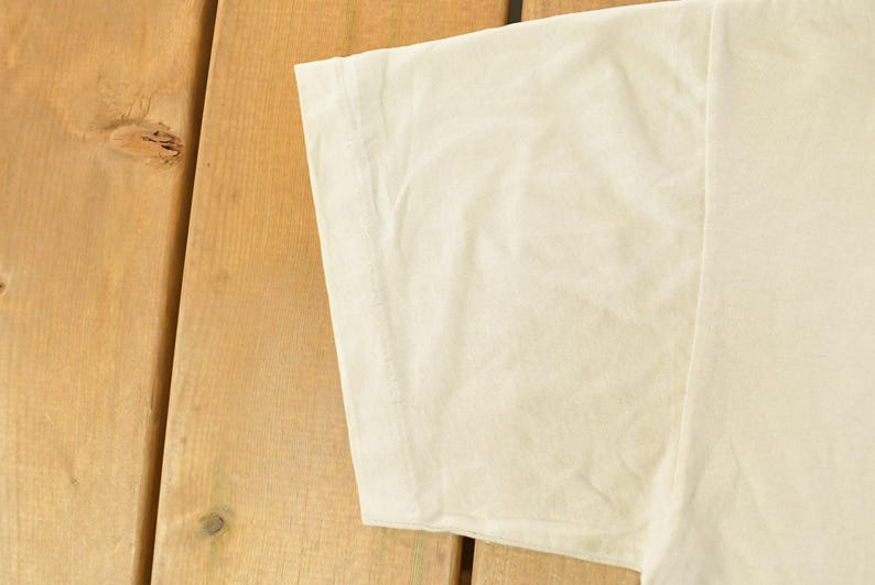 K&ouml;nnte beinhalten: Nahaufnahme eines cremefarbenen T-Shirt-&Auml;rmels. Der Stoff ist leicht zerknittert und der &Auml;rmel ist vor einem Holzgrund platziert. Das Shirt scheint aus einem weichen, leichten Material zu sein.