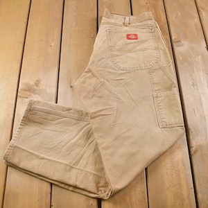 May include: A pair of tan work trousers with a red patch on the back pocket. The trousers are folded and laid out on a wooden surface.