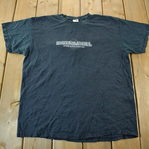 May include: Dark grey t-shirt featuring the white text "CHRISTIANIA" above "GRAND ALLEY COMPLEX". The shirt has a crew neck and is made of a soft material. The t-shirt is laid flat on a wooden surface.