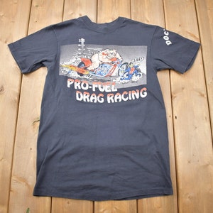 May include: A dark blue t-shirt with a cartoon illustration of a pig riding a motorcycle. The text "PRO-FUEL DRAG RACING" is printed on the front of the shirt.