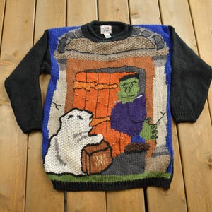 Op de afbeelding: Een blauwe, zwarte en witte gebreide trui met een Halloween-thema. De trui heeft een spook, een monster en een deur met een pompoen erop.