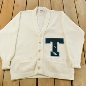 May include: Cream-coloured cardigan sweater with a V-neck and button closure. Features a large, dark green and light blue letter "T" patch on the front. Includes two front pockets and ribbed cuffs. The sweater is displayed on a wooden surface.