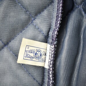 May include: A close-up of a blue quilted fabric with a white tag that reads "GARMENT OF AMERICA" with a logo of a bald eagle holding a shield with the text "UNITED STATES OF AMERICA" inside the shield. The tag also has the letter "C" printed on it.