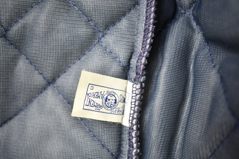 May include: A close-up of a blue quilted fabric with a white tag that reads "GARMENT OF AMERICA" with a logo of a bald eagle holding a shield with the text "UNITED STATES OF AMERICA" inside the shield. The tag also has the letter "C" printed on it.