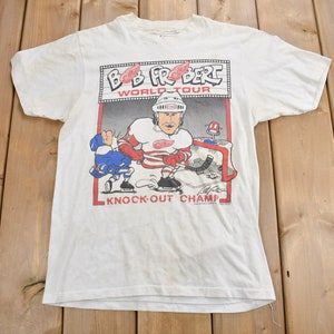 May include: White t-shirt featuring a cartoon image of hockey player Bob Probert on the front. The image is in a red, white and blue colour scheme and includes the text "Bob Probert World Tour Knock-Out Champ".