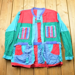 May include: A vintage patchwork jacket in vibrant colours. The jacket has panels of red, teal, green, and blue. It features two chest pockets with vertical striped accents and two lower pockets. The jacket is laid flat on a wooden surface.
