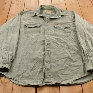May include: A sage green long-sleeve button-down shirt featuring a classic collar, two chest pockets with buttoned flaps, and a full-length front button closure. The garment has long sleeves with buttoned cuffs and a structured, collared neckline.