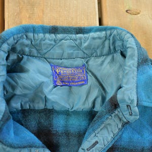May include: Close-up of a blue and black plaid wool shirt. The shirt has a quilted lining and a collar. A blue label with gold text reads "Pendleton Pure Virgin Wool".