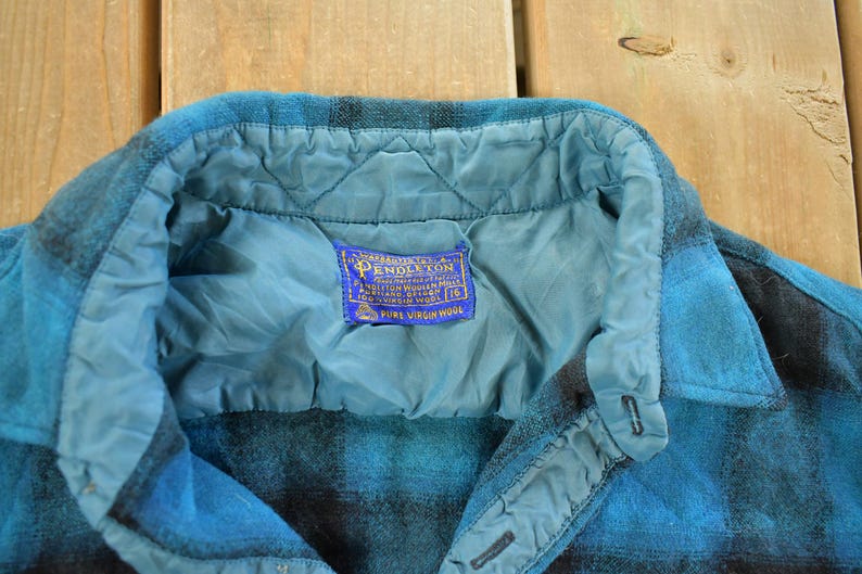 May include: Close-up of a blue and black plaid wool shirt. The shirt has a quilted lining and a collar. A blue label with gold text reads "Pendleton Pure Virgin Wool".
