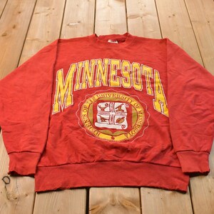 Könnte beinhalten: Ein rotes Langarm-Sweatshirt mit dem Wort MINNESOTA in großen gelben Buchstaben, die über einem kreisförmigen Siegel der University of Minnesota gewölbt sind. Das Siegel enthält eine weiße Schriftrolle mit kleinen Illustrationen und Text.